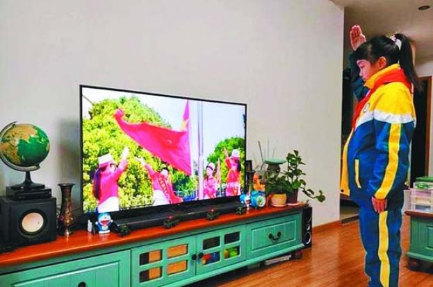学生笔挺地站在家里电视屏幕前，胸口系著红领巾，朝著电视屏幕中的五星红旗敬礼。